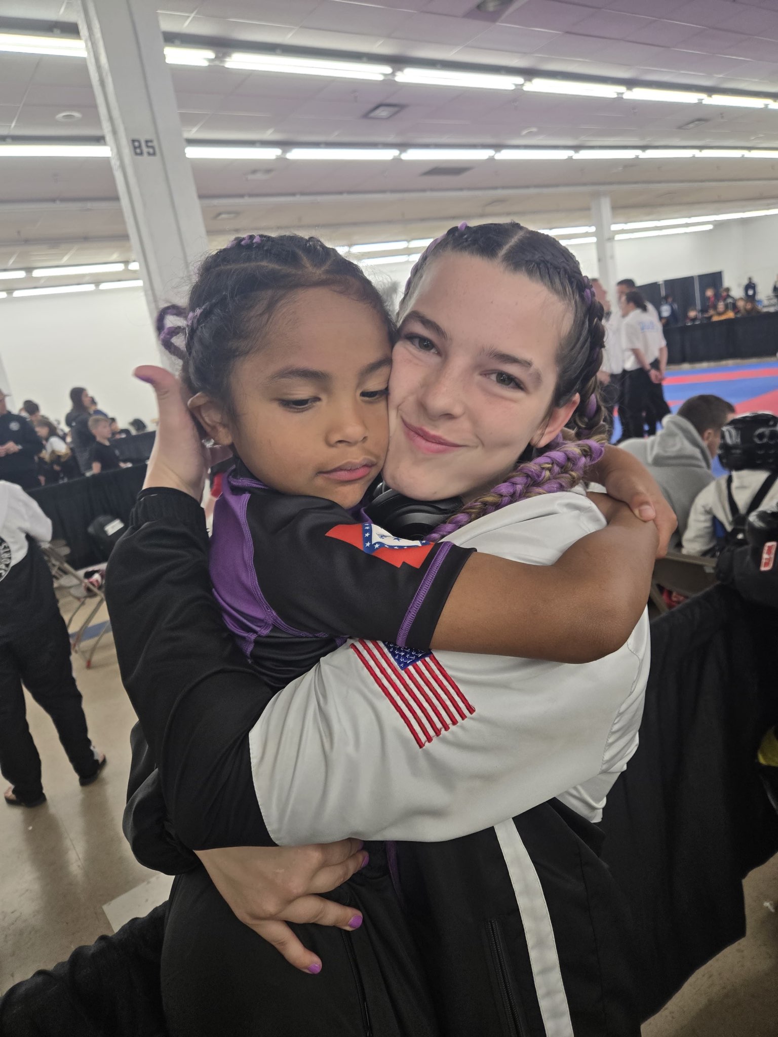 Older TCB female fighter with purple braids hugging a young child in matching purple team jacket at the 2026 WAKO USA Nationals