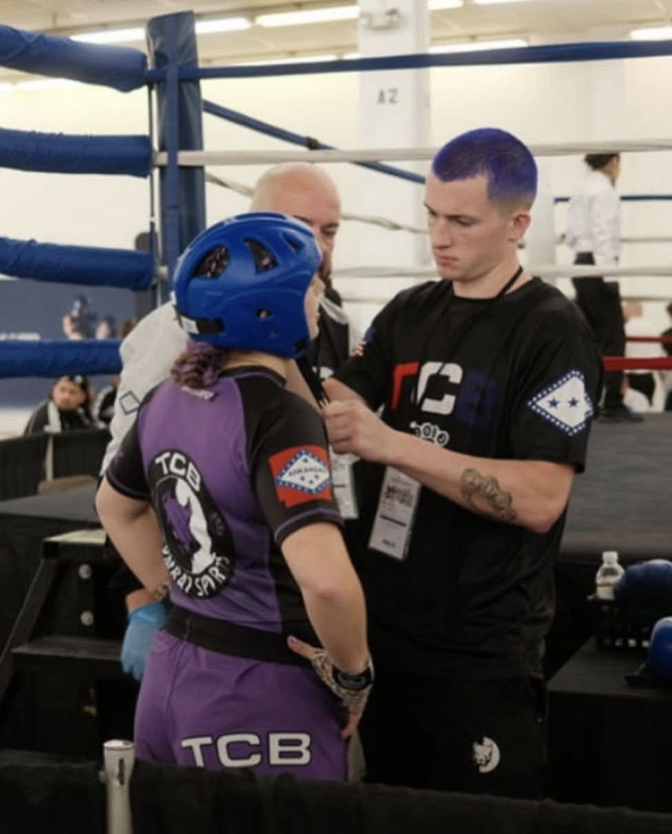 TCB coach taping up a female fighter in purple TCB jersey and blue head guard before her WAKO bout