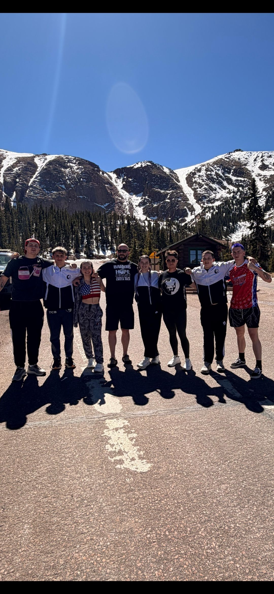 TCB team standing arm-in-arm at a Colorado mountain pass overlook with snowy peaks behind them