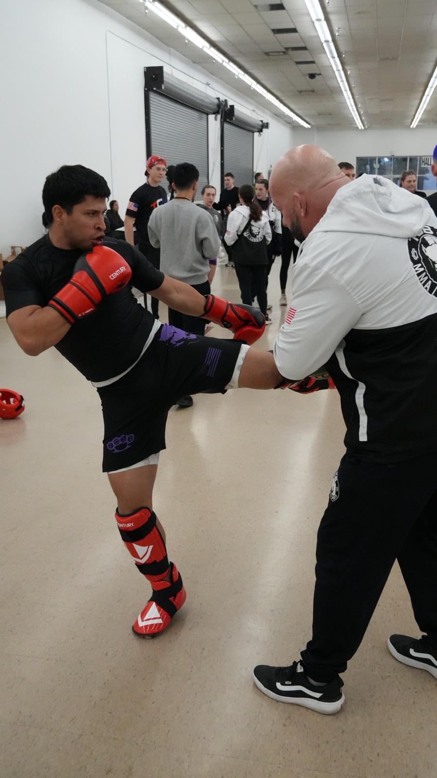 TCB fighter throwing a round kick into a coach's pad in the gym, the rest of the team in the background