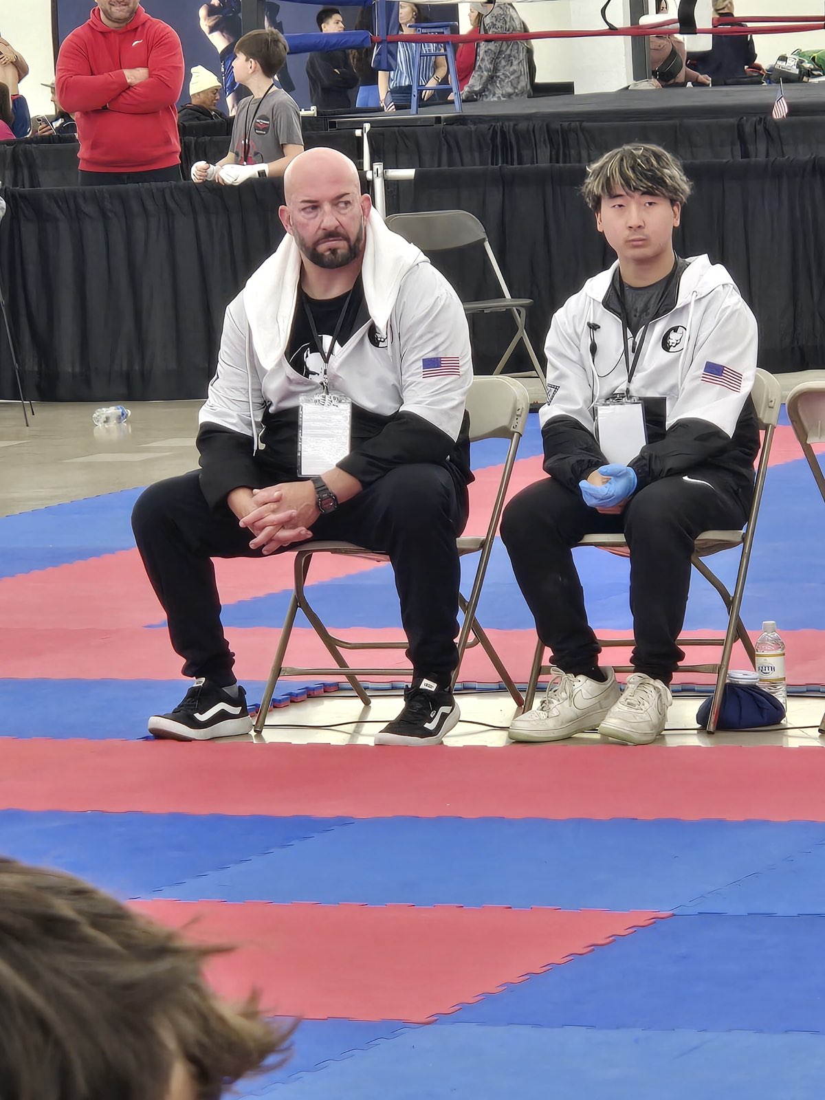 Papa Pit sitting beside a TCB fighter on the venue mats, both watching the action at the 2026 WAKO USA Nationals