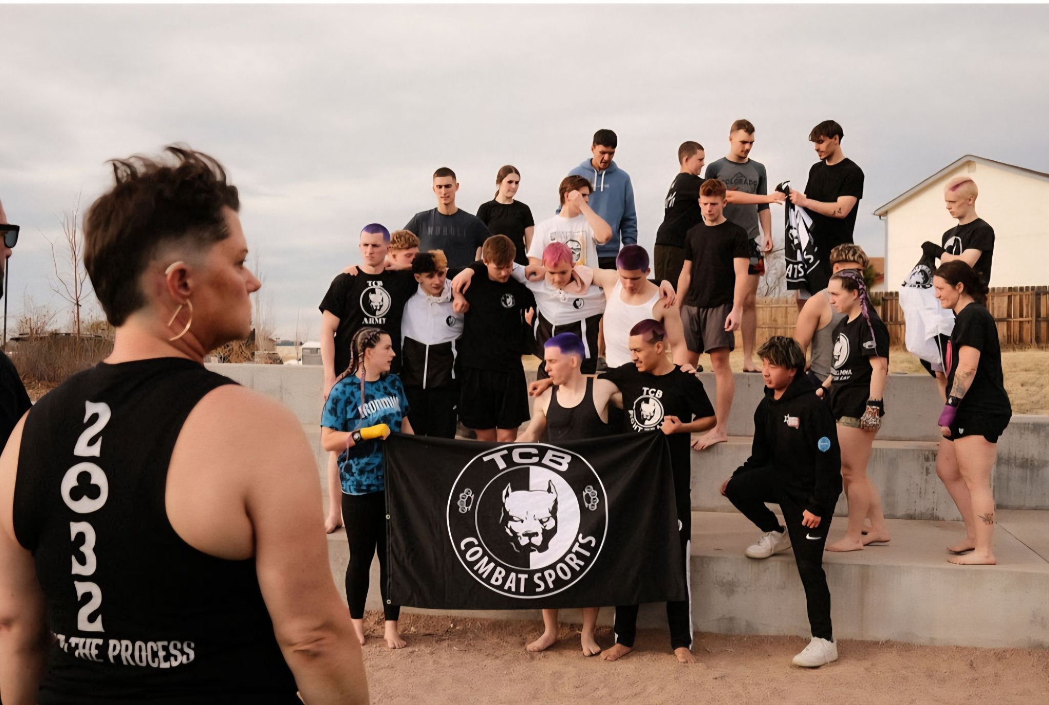 TCB Combat Sports team gathered on the bleachers with the team flag, woman in '2032 Trust The Process' tank top in foreground