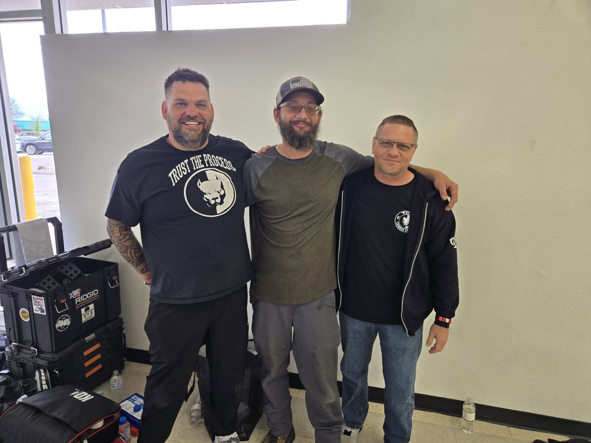 Three TCB Combat Sports men posing arm in arm, the leftmost wearing a 'Trust The Process' pit bull tee