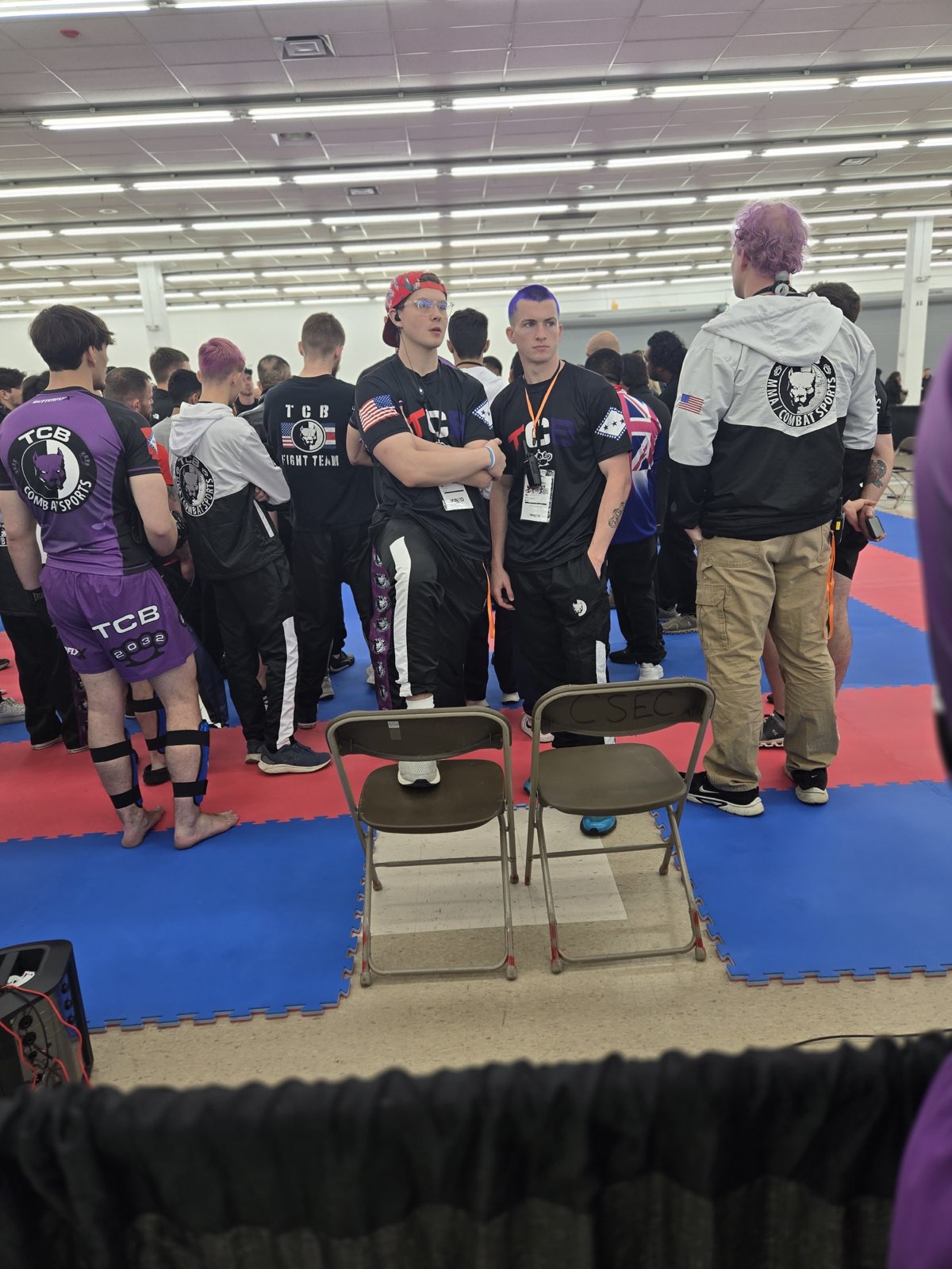 Two TCB fighters in matching team gear waiting in the warm-up area at the WAKO USA venue, one wearing a red bandana, the other with purple hair