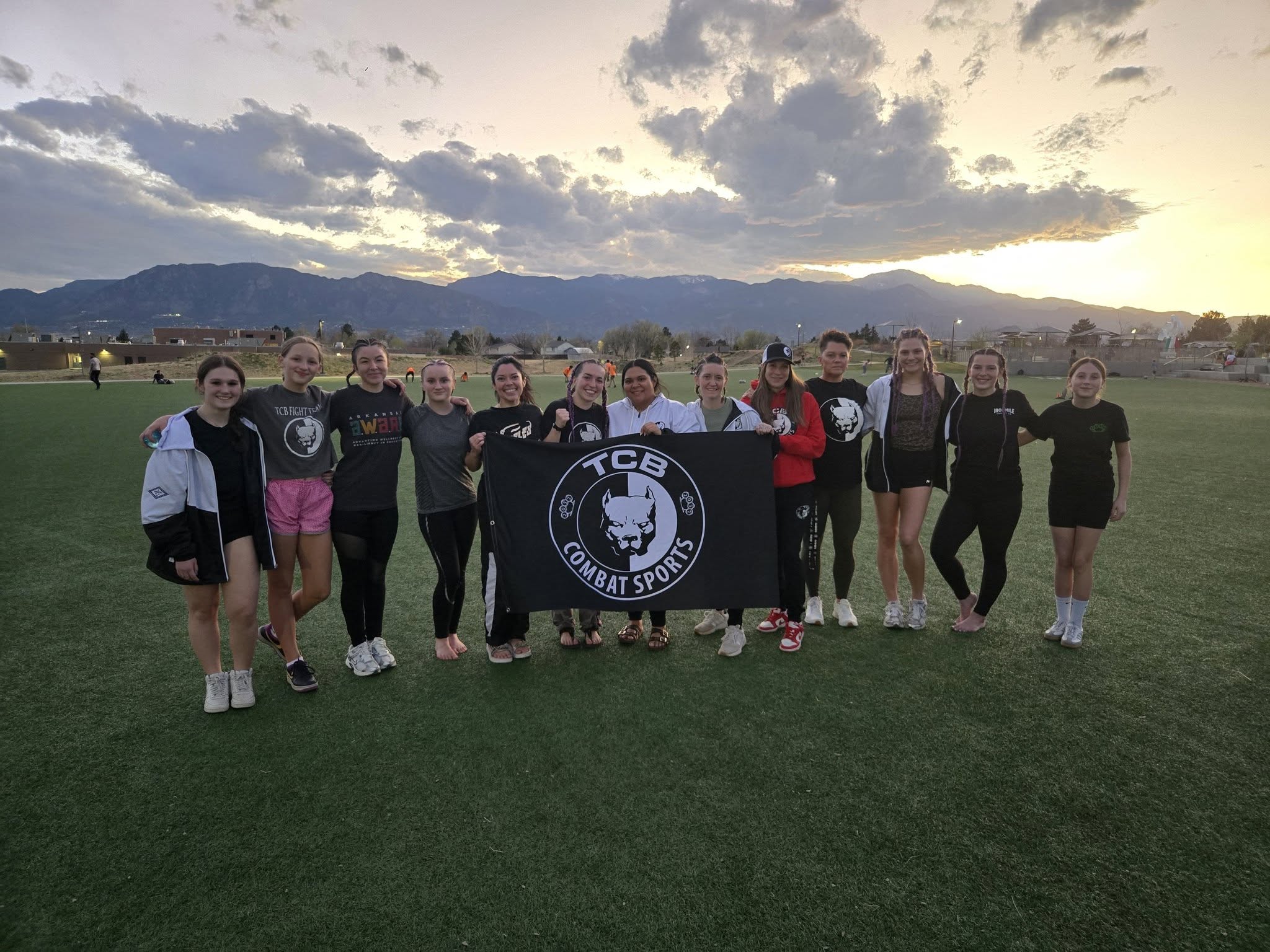 The women of TCB Combat Sports posing with the team flag at sunset in front of the Colorado mountains